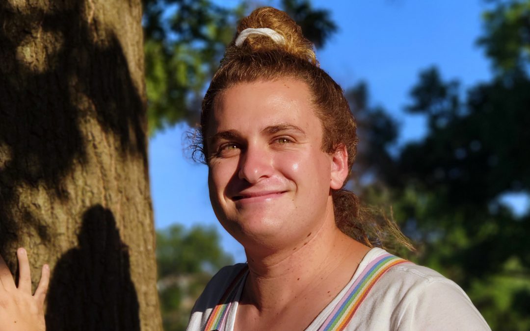 Brienne Hayes wearing pride suspenders poses with hand resting on tree.
