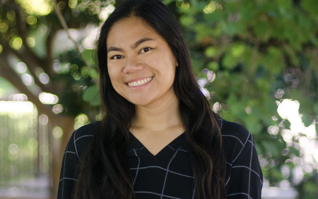 A woman with long dark hair in a long-sleeved black plaid top smiles outside under a tree