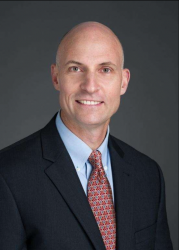 A professional photo of a man in a suit with a red patterned tie in front of a grey background