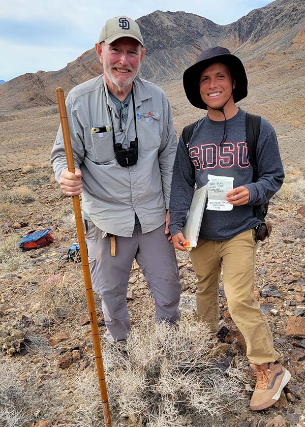 An older man wearing a baseball cap and holding a tall wooden pole stands next to a young male student wearing an SDSU sweatshirt outside. Photo credit: Rob Hawk