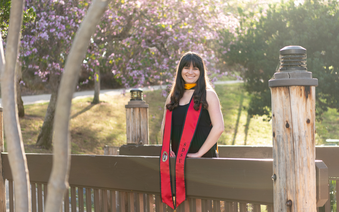 Woman with long dark brown hair wearing a black dress and red graduation sash stands on a bridge during golden hour. A tree with purple blossoms is int he background