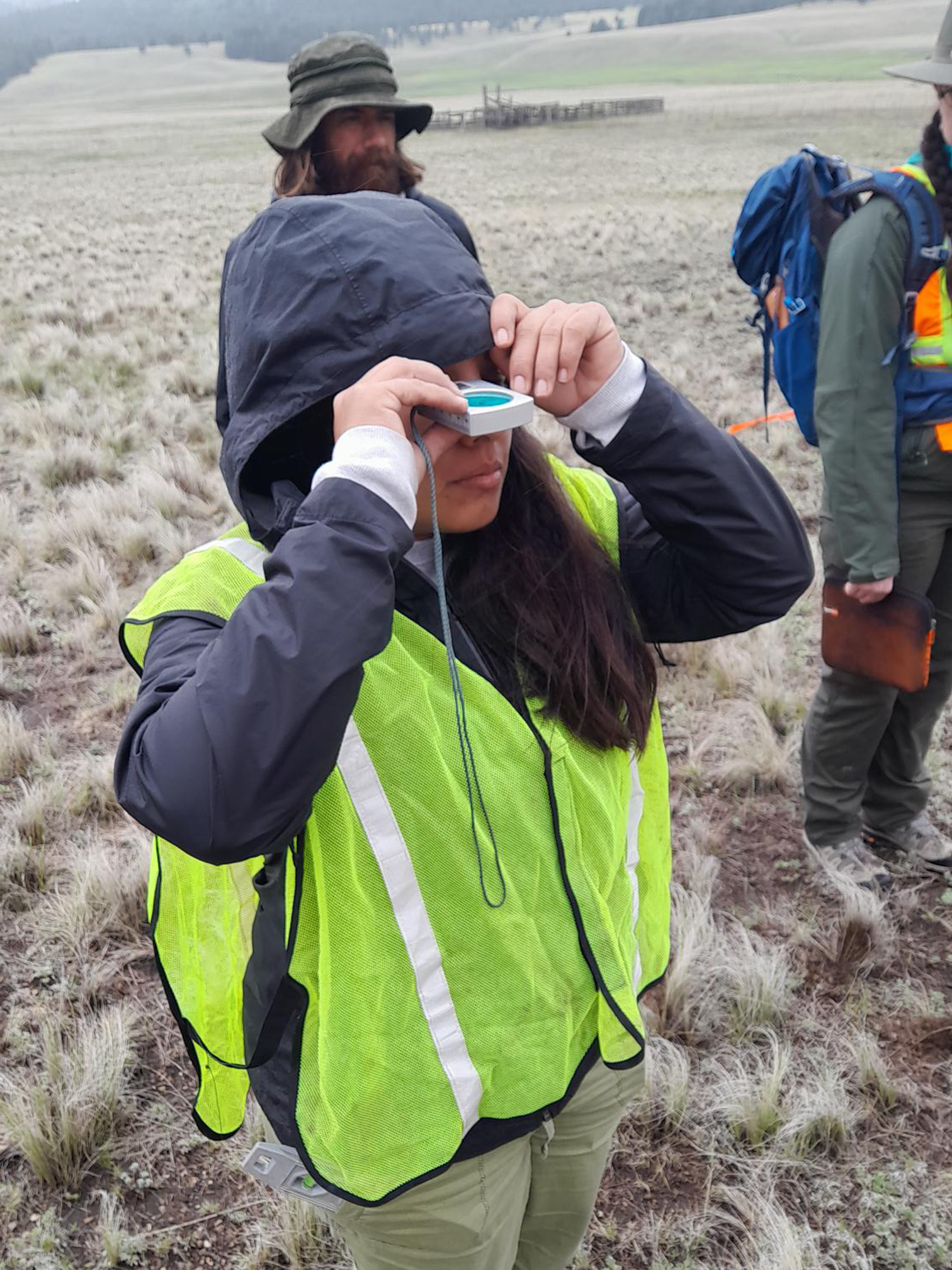 Vanessa, dressed in a neon vest and hoodie, uses a surveying tool in a field in New Mexico