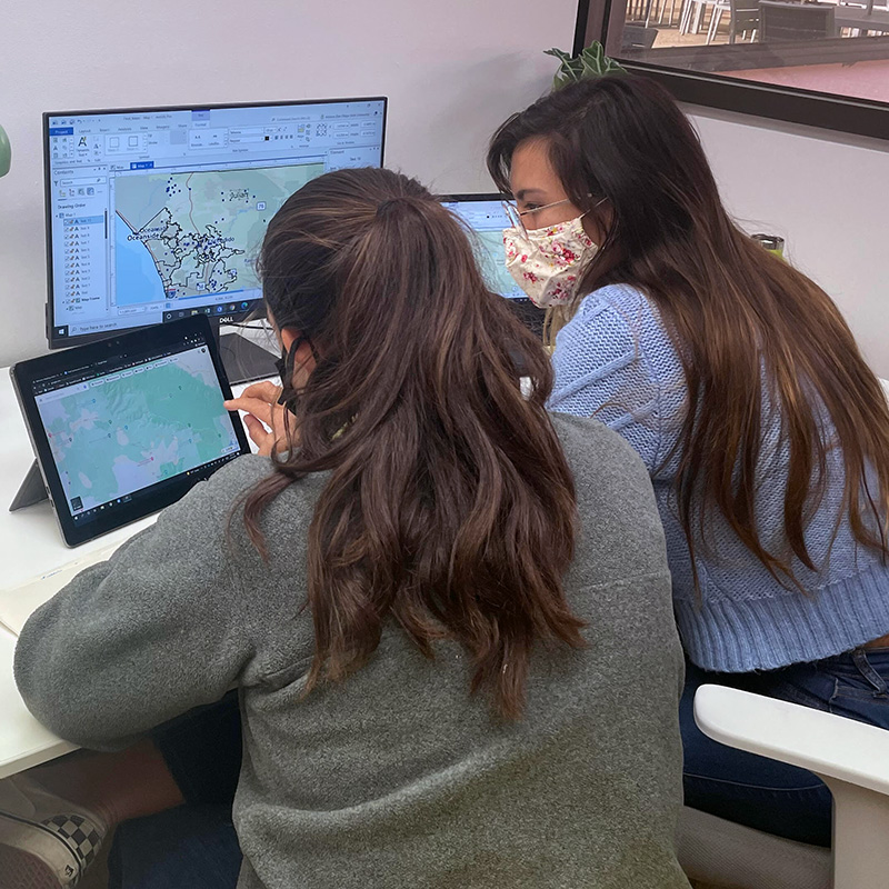 adriana-rios-desktop-maps-800sq Adriana Rios and colleague sitting at desktop facing computer screens with maps.